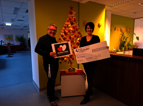Zwei Personen stehen in einem Büro vor einem geschmückten Weihnachtsbaum. Der Mann hält eine Karte mit Herzsymbol, die Frau präsentiert einen großen Scheck – offensichtlich eine Spenden- oder Preisübergabe.