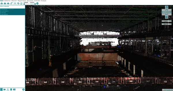 Große Industriehalle unter Bau, Stahlgerüst und Stahlträger umgeben einen dunklen Innenraum. Zentral führt eine tiefe Baugrube bzw. Kanal bis zu einem hell beleuchteten Ausgang am gegenüberliegenden Ende. Rund um die Grube stehen Sicherheitszäune; links und rechts erstrecken sich weitere Gerüste.