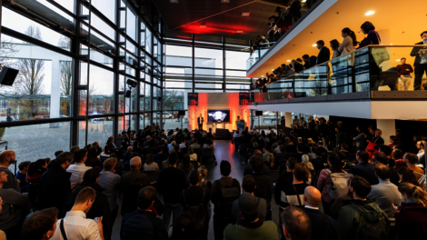 Große Menschenmenge versammelt sich in einem modernen Glas-Atrium zu einer Konferenz. Unten auf der Bühne wird präsentiert, während auf einer oberen Galerie weitere Zuhörer zuschauen. Warme rote Bühnenbeleuchtung und ein Bildschirm mit einer Präsentation im Hintergrund.