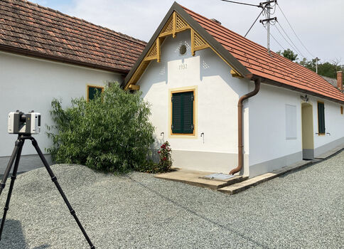 Kleines, weißes Haus mit rotem Ziegeldach und gelbem Giebel. Grüner Fensterladen, Eingang rechts. Eine Pflanze vor der Fassade. Vor dem Gebäude steht ein Kamerastativ, als würde das Haus fotografiert.