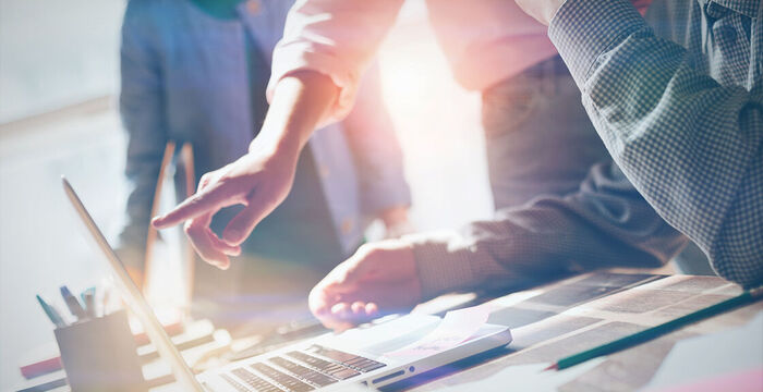 Individuelle-Workshops_Systemeinführung_Headerbild Eine Gruppe von Menschen arbeitet an einem Tisch. Einer zeigt auf das Laptop-Display, während die anderen Notizen und Unterlagen beisteuern. Die Szene vermittelt Zusammenarbeit, Ideenfindung und Fokus in einem hellen Büroraum.