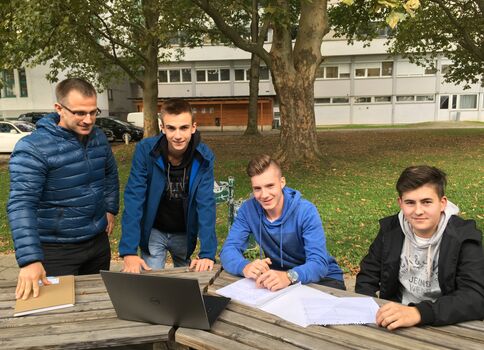 Vier junge Männer arbeiten draußen an einem Laptop und Papieren auf einem Holztisch; im Hintergrund stehen Bäume und ein Gebäude.