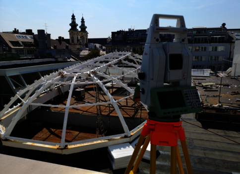 Auf einem Dach steht eine Vermessungsausrüstung (Totale Station) auf einem Stativ. Neben ihr eine weiße geodätische Kuppelrahmenkonstruktion aus Metallbögen. Im Hintergrund Stadtgebäude mit Kirchtürmen, der Himmel ist blau.