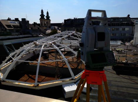 Auf einem Dach steht eine Vermessungsausrüstung (Totale Station) auf einem Stativ. Neben ihr eine weiße geodätische Kuppelrahmenkonstruktion aus Metallbögen. Im Hintergrund Stadtgebäude mit Kirchtürmen, der Himmel ist blau.