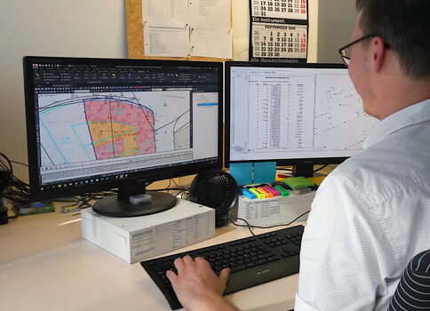 Ein Mann sitzt an einem Schreibtisch mit zwei Monitoren. Links ist eine GIS-Karte mit farblich markierten Zonen zu sehen, rechts eine Datentabelle. Er tippt auf der Tastatur und analysiert Karten und Daten.