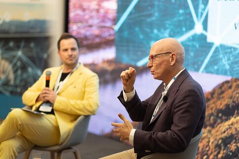 Zwei Männer führen ein Panelgespräch auf einer Bühne. Der linke Mann in gelbem Anzug hält ein Mikrofon; der rechte Mann in dunklem Sakko und Brille spricht mit Handgesten. Im Hintergrund sind digitale Grafiken zu sehen.