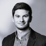 Schwarzweißes Porträt eines jungen Mannes in Blazer und Hemd, frontal zur Kamera, mit leichtem Lächeln; neutraler Hintergrund.