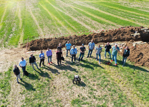 Eine Gruppe von etwa 15 Personen steht im Kreis auf einem Feld um eine abgegrenzte Grube; rotes Absperrband markiert den Bereich. In der Mitte liegt ein kleines, dunkles Gerät, vermutlich eine Demonstrationsausrüstung.