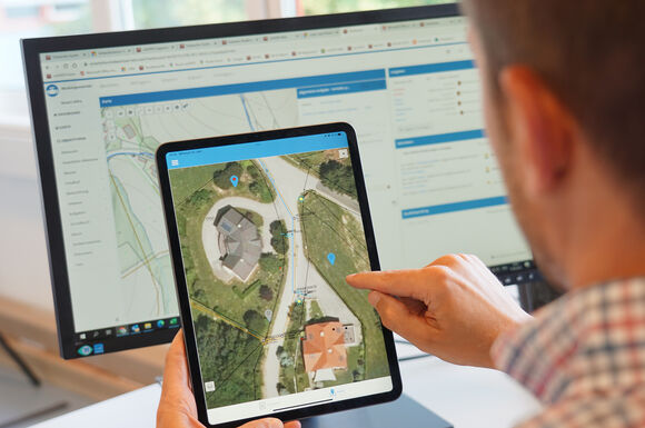 Eine Person hält ein Tablet mit einer Satellitenkartenansicht eines Gebäudekomplexes; auf der Karte sind Gebäude, Wege und Standortmarker sichtbar, während im Hintergrund ein Computerbildschirm eine ähnliche Kartenansicht zeigt.