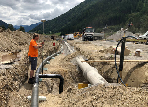 Auf einer Baustelle in einer bergigen Landschaft arbeitet ein Mann in Orange an einer großen schwarzen Rohrleitung, die durch einen Graben verläuft. Im Hintergrund stehen Baufahrzeuge, Berge und Wolken am Himmel.