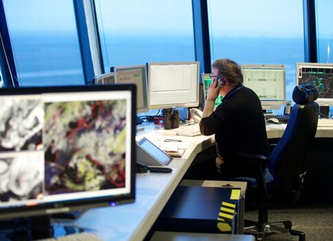 Eine Frau arbeitet in einem Kontrollraum, umgeben von mehreren Monitoren mit Diagrammen und Karten. Sie sitzt an einem Schreibtisch, telefoniert und blickt konzentriert auf den Bildschirm. Große Fenster geben einen Blick aufs Meer und den Himmel frei.