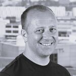 Porträt eines lächelnden Mannes mittleren Alters in einem schwarzen T-Shirt. Graustufenbild vor städtischem Hintergrund mit Gebäuden. Der Mann blickt leicht zur Seite und wirkt freundlich.