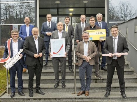 Eine Gruppe Geschäftsleute in Anzügen steht auf einer Treppe vor einem Bürogebäude. Einige halten große Schilder mit Hashtags, ein Mann präsentiert ein gerahmtes Zertifikat mit rotem Häkchen, ein weiterer einen Scheck über 5.000 Euro. Feierstimmung bei einer Preisverleihung.