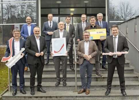 Eine Gruppe Geschäftsleute in Anzügen steht auf einer Treppe vor einem Bürogebäude. Einige halten große Schilder mit Hashtags, ein Mann präsentiert ein gerahmtes Zertifikat mit rotem Häkchen, ein weiterer einen Scheck über 5.000 Euro. Feierstimmung bei einer Preisverleihung.