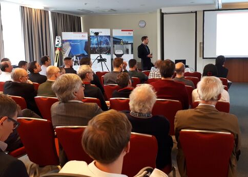 Eine Präsentation vor Seminarpublikum: Ein Redner steht vorne neben Beamer und Leinwand, während zahlreiche Zuhörer in roten Sesseln dem Vortrag folgen.