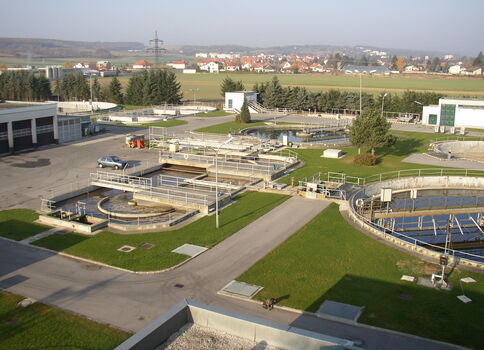 Luftaufnahme eines Klärwerks: mehrere runde Becken mit Rohrleitungen, angrenzende Wege und Grünflächen; im Hintergrund eine kleine Stadt mit Häusern.
