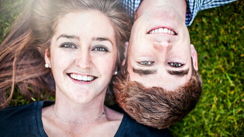 Ein lächelndes Paar liegt gemeinsam auf dem Gras, Kopf an Kopf, und schaut in die Kamera.