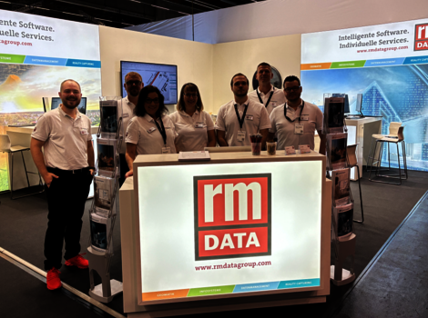 Gruppe von sieben Mitarbeitenden in weißen Poloshirts steht hinter einem leuchtenden Empfangstresen mit dem RM DATA-Logo; im Hintergrund Messestand mit Bannern.