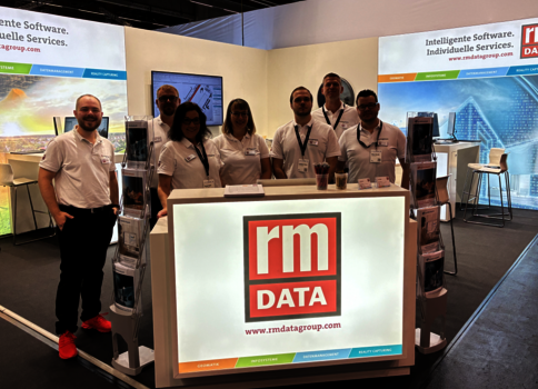 Gruppe von sieben Mitarbeitenden in weißen Poloshirts steht hinter einem leuchtenden Empfangstresen mit dem RM DATA-Logo; im Hintergrund Messestand mit Bannern.
