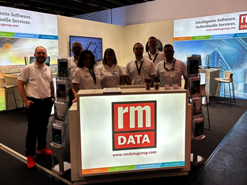 Gruppe von sieben Mitarbeitenden in weißen Poloshirts steht hinter einem leuchtenden Empfangstresen mit dem RM DATA-Logo; im Hintergrund Messestand mit Bannern.