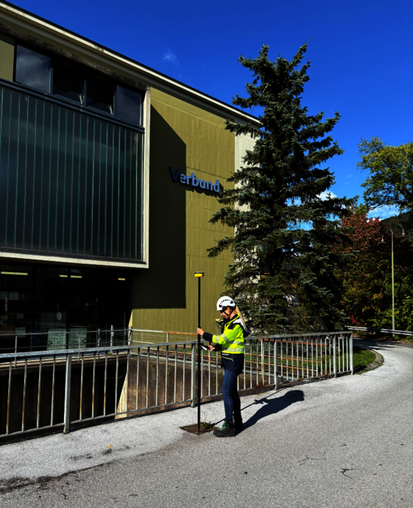 Eine Sicherheitskraft in gelber Warnweste und Helm steht am Gehweg neben einem Metallgeländer und hält einen langen Stab. Dahinter befindet sich ein modernes Gebäude mit der Aufschrift 'Verbund'; sonniger Tag, blauer Himmel und ein großer Baum im Hintergrund.