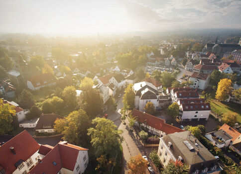 Luftaufnahme einer idyllischen Kleinstadt von oben: rote Ziegeldächer, grüne Bäume, enge Straßen und ein Kirchturm am Horizont; sanftes Sonnenlicht über der Szene.