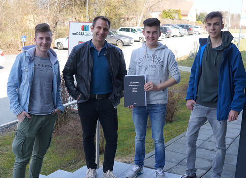 Vier junge Männer stehen draußen nebeneinander auf einer Treppe. Einer hält ein graues Dokument, die anderen lächeln zur Kamera. Im Hintergrund sind Autos und Bäume sichtbar.
