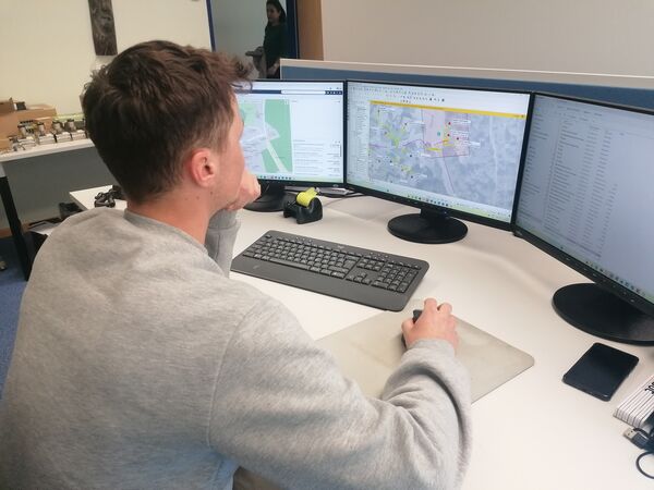 Ein Mann sitzt an einem Schreibtisch vor drei Monitoren, auf denen Karten und Diagramme angezeigt werden. Er tippt an der Tastatur, eine Maus liegt daneben; Notizblock und Smartphone befinden sich auf dem Tisch, im Büro steht weiteres Equipment.