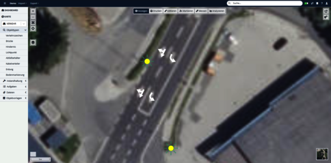 Satellitenansicht einer mehrspurigen Straße mit Kreuzung, Gebäuden am Rand und zwei gelben Markern auf der Fahrbahn, vermutlich Wegpunkte in einer Kartensoftware.