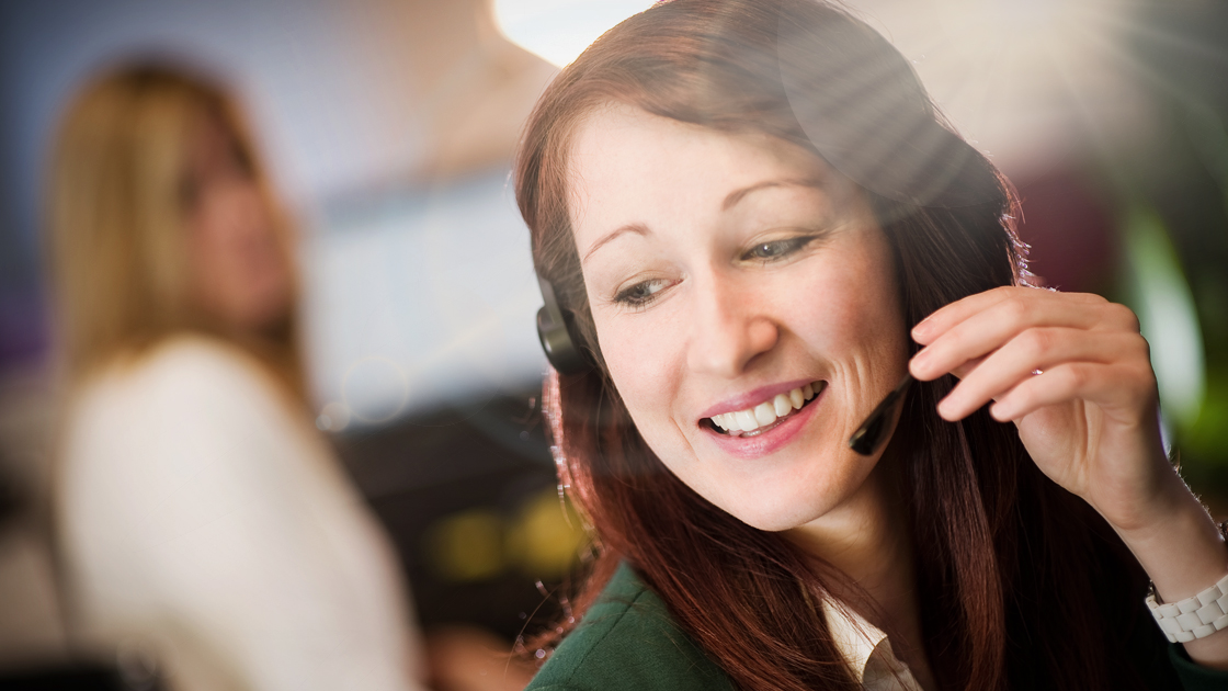 Eine junge Frau mit Headset lächelt freundlich; sie arbeitet vermutlich im Kundenservice in einem Büro, im Hintergrund ist eine weitere Person unscharf zu sehen.